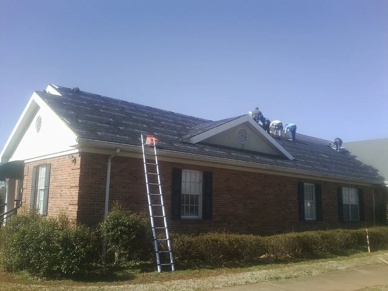 Crew tearing shingles off a brick ranch roof with a ladder set — Emergency Roof Repair work by our crew in Hickory Corners