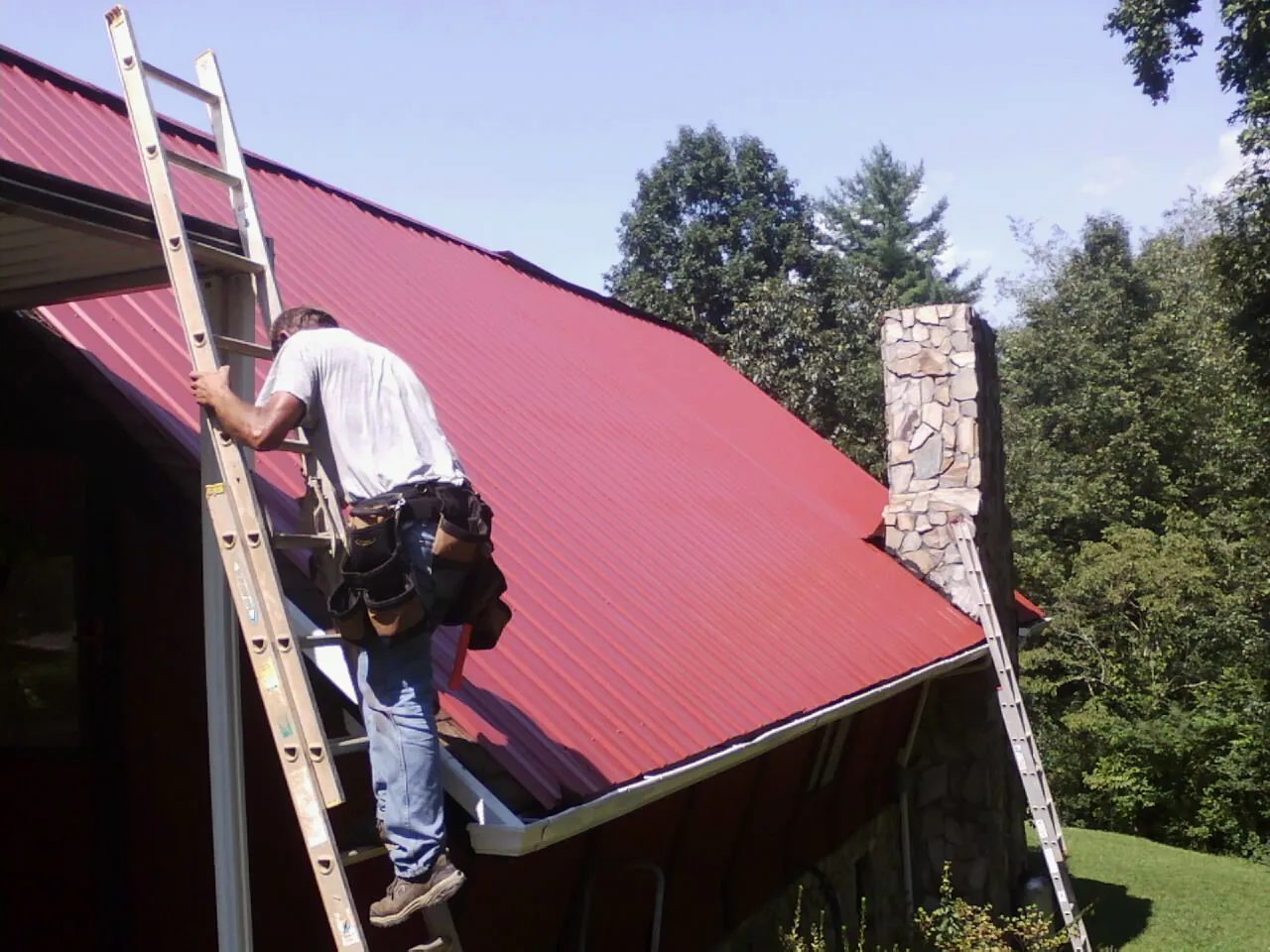 Roofer on a ladder beside a red metal roof and stone chimney — Soffit & Fascia Repair by Roofing Brothers in Hickory Corners